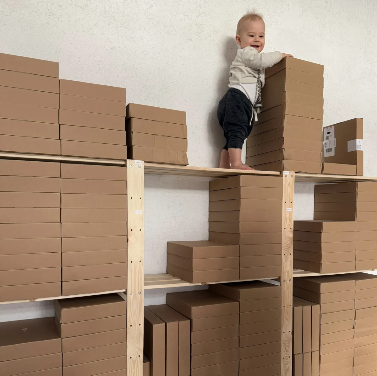 Ein freudiges Kleinkind steht bei Mriella neben ordentlich gestapelten Versandkartons der Babyalben – passend zum Slogan "Ein Leben. Box für Box."